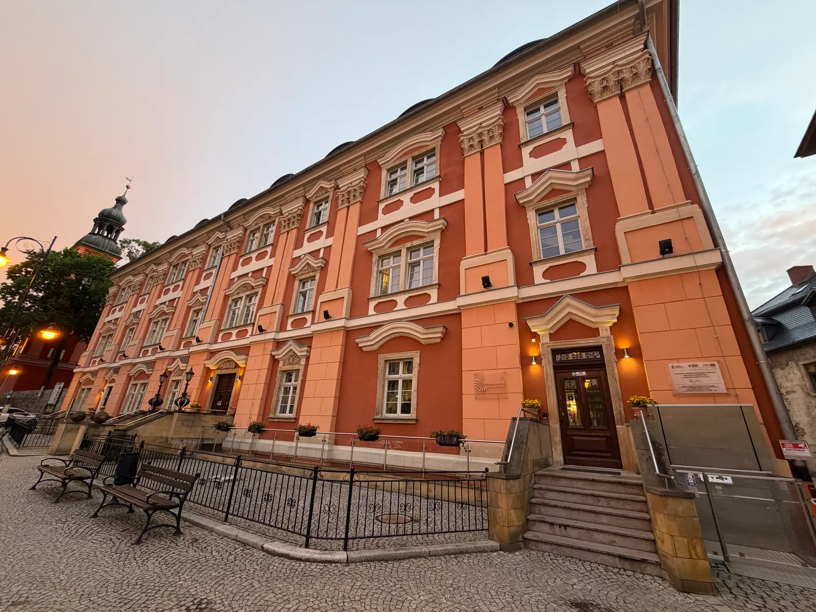 Długi Dom w Cieplicach o zmierzchu – wydłużona, trzykondygnacyjna elewacja w kolorze łososiowym, po lewej wieża kościoła świętego Jana Chrzciciela