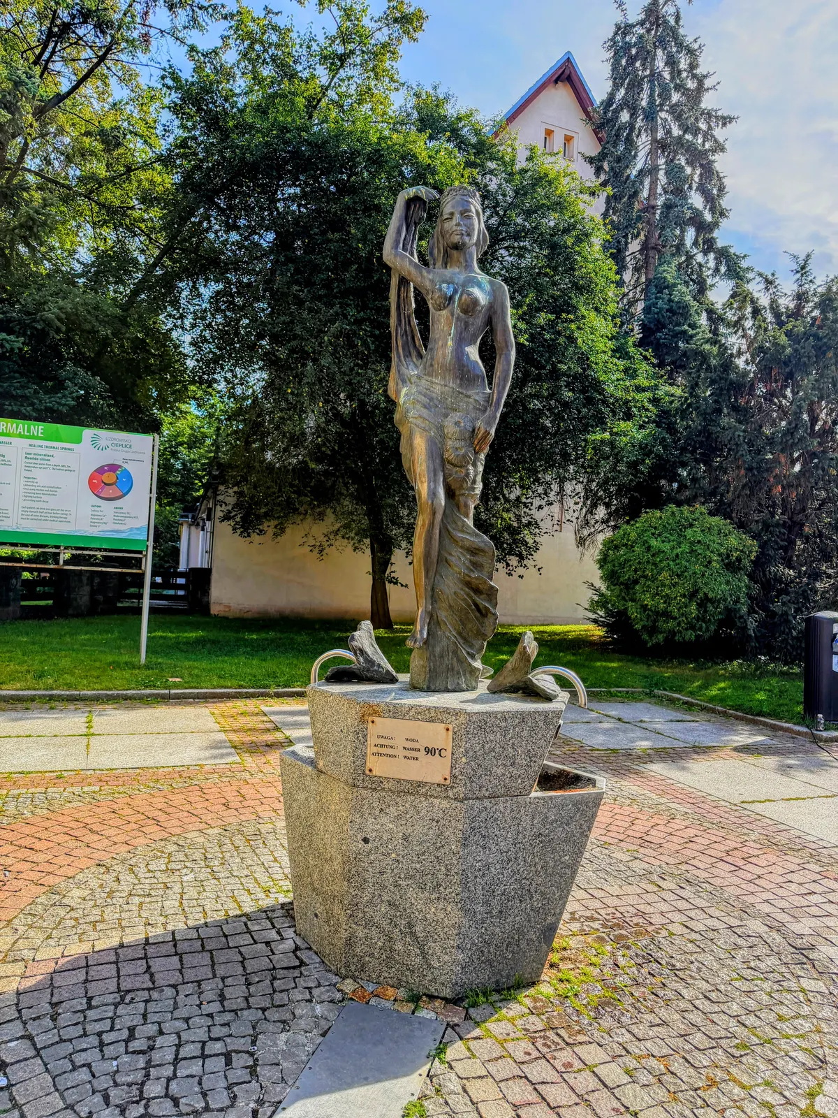 Brązowa rzeźba nimfy wodnej na granitowym cokole fontanny w Cieplicach; na cokole tabliczka „UWAGA! WODA 90°C", po lewej tablica informacyjna Uzdrowiska Cieplice o leczniczych wodach termalnych
