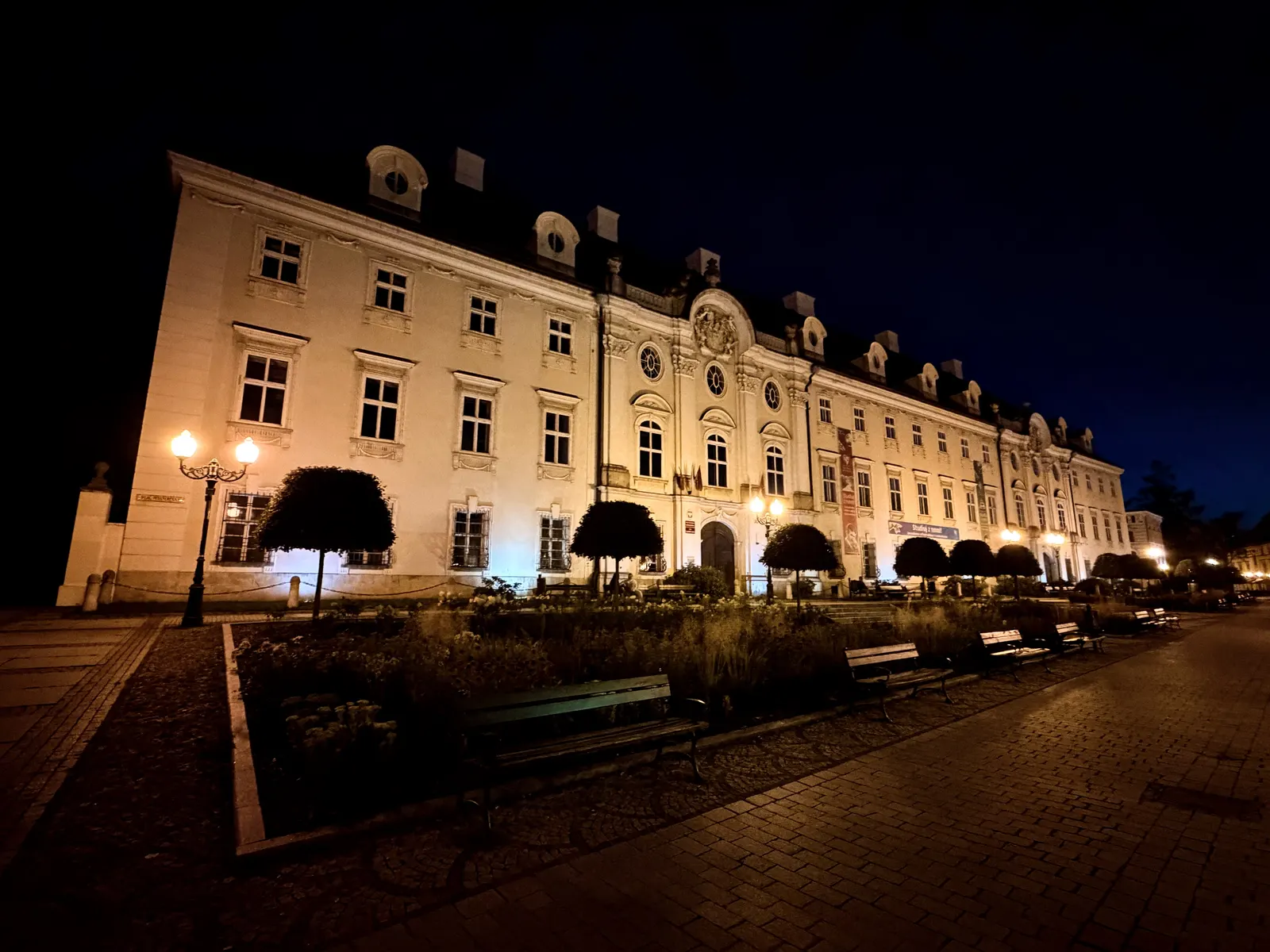 Podświetlona nocą fasada pałacu Schaffgotschów w Cieplicach widziana z placu Piastowskiego: długa, trzykondygnacyjna elewacja z ryzalitem zwieńczonym kartuszem herbowym, owalne lukarny w dachu mansardowym, przed budynkiem strzyżone drzewa i ławki