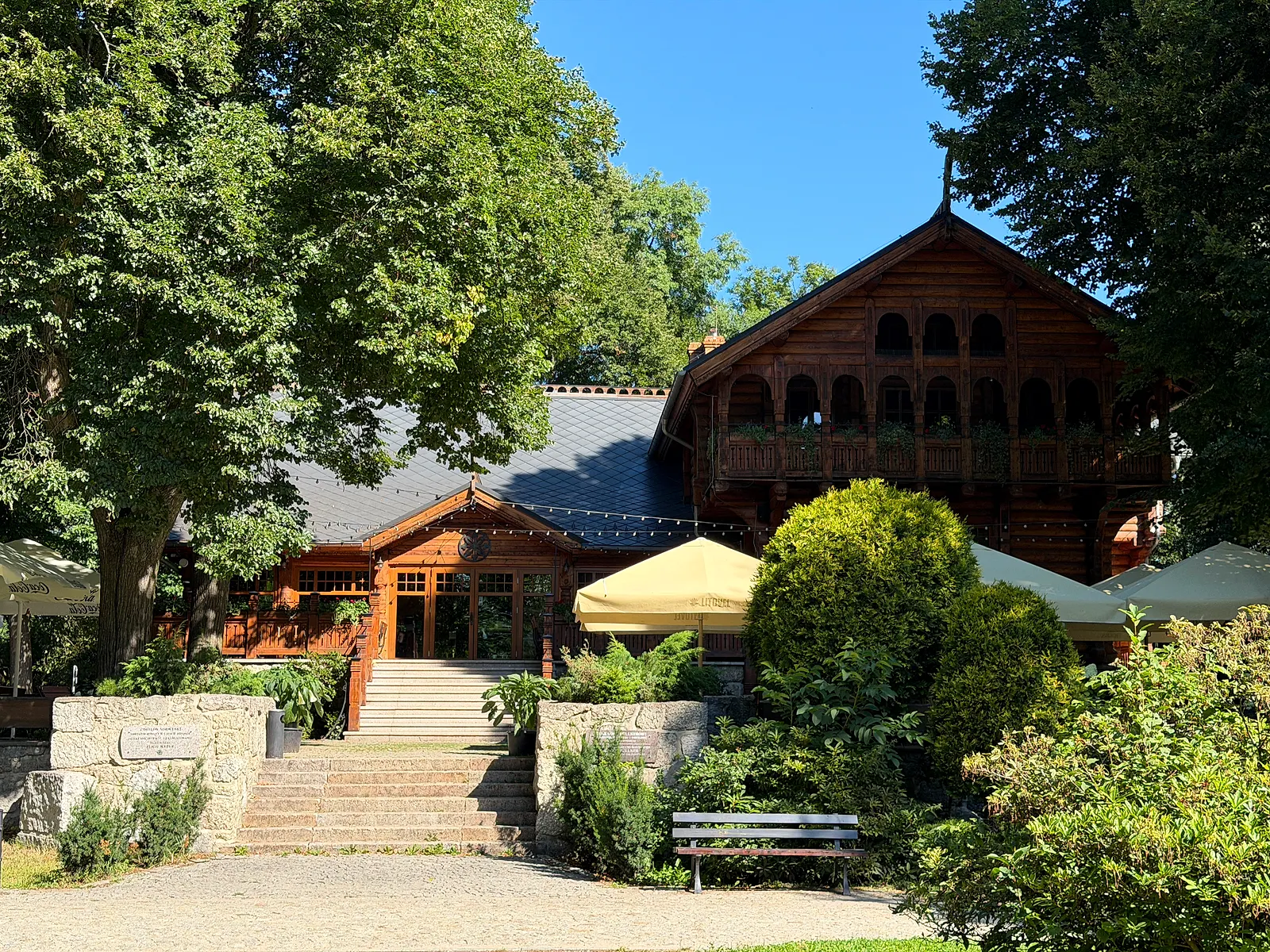 Pawilon Norweski w Parku Norweskim w Cieplicach: ciemna drewniana bryła ze zdobionym szczytem i galeryjką na piętrze, niżej przeszklona weranda, przed wejściem kamienne schody, parasole ogródka i ławka